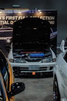 A modified white car is on display at an indoor automotive show, with the hood open to reveal a blue engine compartment. The setup includes a backdrop with partially visible signage and lighting fixtures. Nearby vehicles are partially visible, with one orange car in the foreground. The license plate features Japanese characters.