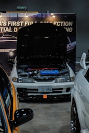 A modified white car is on display at an indoor automotive show, with the hood open to reveal a blue engine compartment. The setup includes a backdrop with partially visible signage and lighting fixtures. Nearby vehicles are partially visible, with one orange car in the foreground. The license plate features Japanese characters.