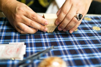 A person is folding a piece of paper on a blue plaid tablecloth. The hands are adorned with a ring. Various items can be seen on the table, including a small block and what appears to be packets of paper.