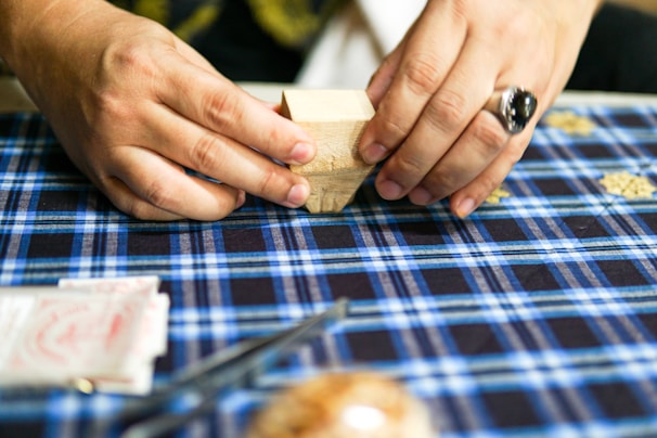A person is folding a piece of paper on a blue plaid tablecloth. The hands are adorned with a ring. Various items can be seen on the table, including a small block and what appears to be packets of paper.