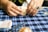 A person is folding a piece of paper on a blue plaid tablecloth. The hands are adorned with a ring. Various items can be seen on the table, including a small block and what appears to be packets of paper.