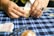 A person is folding a piece of paper on a blue plaid tablecloth. The hands are adorned with a ring. Various items can be seen on the table, including a small block and what appears to be packets of paper.