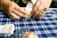 A person is folding a piece of paper on a blue plaid tablecloth. The hands are adorned with a ring. Various items can be seen on the table, including a small block and what appears to be packets of paper.