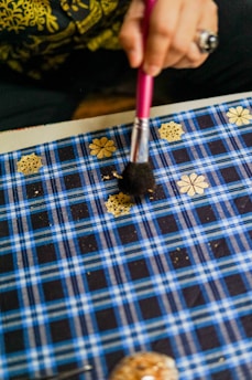 A person is using a brush to apply gold flower and snowflake designs onto a blue plaid fabric. The person's hand and brush are clearly visible along with some intricate details in the background.