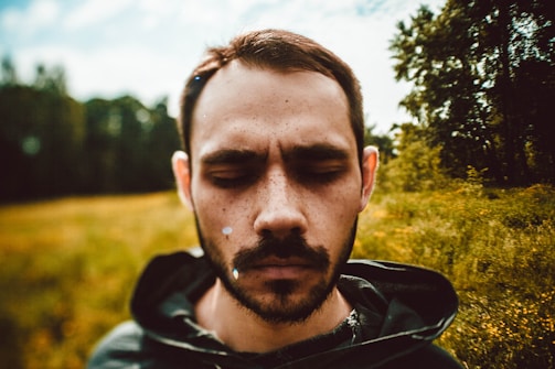 A person with closed eyes stands in an outdoor setting. The background shows a blurred view of trees and grass, suggesting a natural environment with a rustic vibe. The individual appears contemplative or introspective, with sunlight creating a soft, warm tone.