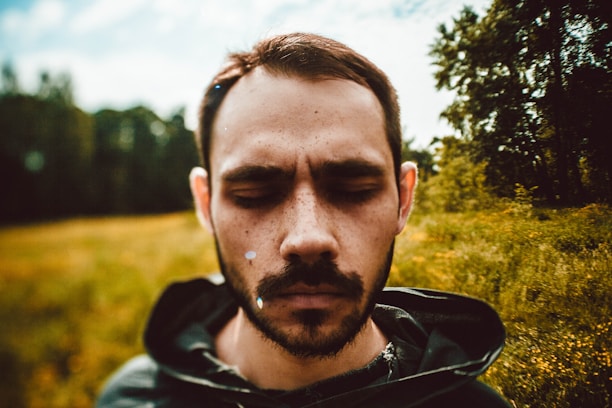 A person with closed eyes stands in an outdoor setting. The background shows a blurred view of trees and grass, suggesting a natural environment with a rustic vibe. The individual appears contemplative or introspective, with sunlight creating a soft, warm tone.