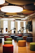 A modern music room with various drums and musical instruments set on a stage. The space features colorful, round seating stools scattered across a wooden floor, and circular acoustic panels hanging from the ceiling. Natural light filters through large windows in the background.