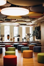 A modern music room with various drums and musical instruments set on a stage. The space features colorful, round seating stools scattered across a wooden floor, and circular acoustic panels hanging from the ceiling. Natural light filters through large windows in the background.