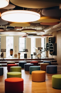 A modern music room with various drums and musical instruments set on a stage. The space features colorful, round seating stools scattered across a wooden floor, and circular acoustic panels hanging from the ceiling. Natural light filters through large windows in the background.