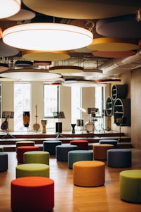 A modern music room with various drums and musical instruments set on a stage. The space features colorful, round seating stools scattered across a wooden floor, and circular acoustic panels hanging from the ceiling. Natural light filters through large windows in the background.