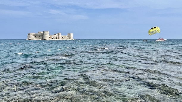 A tranquil seaside scene with a large stone fortification resting on an island, surrounded by clear blue water. A parasail, towed by a speedboat, adds a touch of adventure to the peaceful setting. The sky is expansive and light blue with few clouds.
