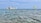 A tranquil seaside scene with a large stone fortification resting on an island, surrounded by clear blue water. A parasail, towed by a speedboat, adds a touch of adventure to the peaceful setting. The sky is expansive and light blue with few clouds.