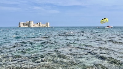 A tranquil seaside scene with a large stone fortification resting on an island, surrounded by clear blue water. A parasail, towed by a speedboat, adds a touch of adventure to the peaceful setting. The sky is expansive and light blue with few clouds.