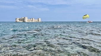 A tranquil seaside scene with a large stone fortification resting on an island, surrounded by clear blue water. A parasail, towed by a speedboat, adds a touch of adventure to the peaceful setting. The sky is expansive and light blue with few clouds.
