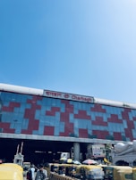 A large building with a modern facade made of glass and red patterns. The name 'Charbagh' is displayed prominently in both English and another script. There are multiple yellow and green auto-rickshaws parked in front of the building. The sky is clear and blue.