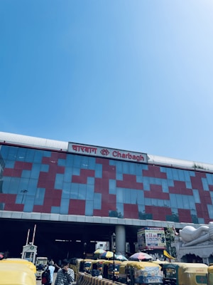 A large building with a modern facade made of glass and red patterns. The name 'Charbagh' is displayed prominently in both English and another script. There are multiple yellow and green auto-rickshaws parked in front of the building. The sky is clear and blue.