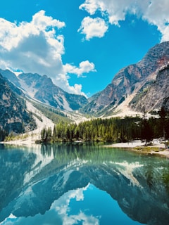A stunning alpine landscape featuring towering mountains with hints of snow, their rugged forms mirrored in a tranquil, clear lake. Pine trees line the shore, and fluffy white clouds drift across a vivid blue sky.