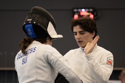 Two fencers wearing protective gear are engaged in a bout. One fencer has their back to the camera, with the name 'Delimatsis' and 'SUI' visible on their jacket. The other fencer, facing the camera, is raising a hand as if in a defensive or conversational gesture. A scoreboard in the background displays red illuminated numbers.