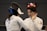 Two fencers wearing protective gear are engaged in a bout. One fencer has their back to the camera, with the name 'Delimatsis' and 'SUI' visible on their jacket. The other fencer, facing the camera, is raising a hand as if in a defensive or conversational gesture. A scoreboard in the background displays red illuminated numbers.