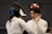 Two fencers wearing protective gear are engaged in a bout. One fencer has their back to the camera, with the name 'Delimatsis' and 'SUI' visible on their jacket. The other fencer, facing the camera, is raising a hand as if in a defensive or conversational gesture. A scoreboard in the background displays red illuminated numbers.