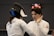 Two fencers wearing protective gear are engaged in a bout. One fencer has their back to the camera, with the name 'Delimatsis' and 'SUI' visible on their jacket. The other fencer, facing the camera, is raising a hand as if in a defensive or conversational gesture. A scoreboard in the background displays red illuminated numbers.