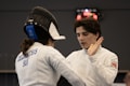 Two fencers wearing protective gear are engaged in a bout. One fencer has their back to the camera, with the name 'Delimatsis' and 'SUI' visible on their jacket. The other fencer, facing the camera, is raising a hand as if in a defensive or conversational gesture. A scoreboard in the background displays red illuminated numbers.
