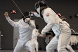 Two fencers in full protective gear, including masks and suits, are engaged in a match. One fencer lunges forward with a foil towards the other, who is in a defensive posture. The background indicates a fencing venue or training area.