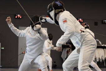 Two fencers in full protective gear, including masks and suits, are engaged in a match. One fencer lunges forward with a foil towards the other, who is in a defensive posture. The background indicates a fencing venue or training area.