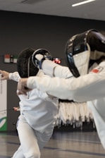 Two fencers are engaged in a duel. They are wearing protective gear, including masks and white uniforms. One fencer is lunging forward with a sword, while the other appears to be attempting a defensive maneuver. The background shows racks with additional fencing attire, indicating an indoor sports facility.