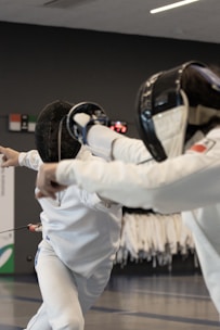 Two fencers are engaged in a duel. They are wearing protective gear, including masks and white uniforms. One fencer is lunging forward with a sword, while the other appears to be attempting a defensive maneuver. The background shows racks with additional fencing attire, indicating an indoor sports facility.