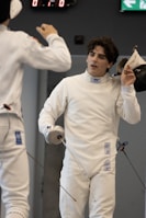 Two fencers in white protective gear are in a duel. One fencer is holding a foil and removing his mask, while the other fencer, also holding a foil, is partially visible. The background shows a digital scoreboard and an exit sign.