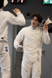 Two fencers in white protective gear are in a duel. One fencer is holding a foil and removing his mask, while the other fencer, also holding a foil, is partially visible. The background shows a digital scoreboard and an exit sign.
