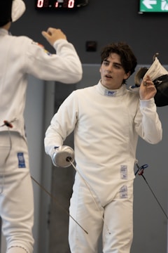Two fencers in white protective gear are in a duel. One fencer is holding a foil and removing his mask, while the other fencer, also holding a foil, is partially visible. The background shows a digital scoreboard and an exit sign.