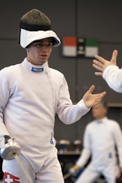A fencer in full white fencing attire stands with a foil in hand, showcasing a focused expression. The fencer wears a mask on top of the head and is engaged in conversation or receiving instruction, with another person partially visible reaching out a hand. A blurred figure in similar attire is in the background.