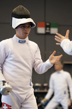 A dynamic fencing coach guiding young athletes during a training session in a bright, modern sports hall.