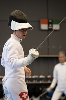 A fencer is dressed in full protective gear, including a mask, jacket, and glove, holding a foil sword in a ready stance. The white uniform has a Swiss flag emblem on the thigh. Another fencer can be seen in the background, slightly blurred.