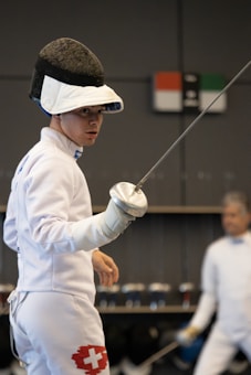 A fencer is dressed in full protective gear, including a mask, jacket, and glove, holding a foil sword in a ready stance. The white uniform has a Swiss flag emblem on the thigh. Another fencer can be seen in the background, slightly blurred.