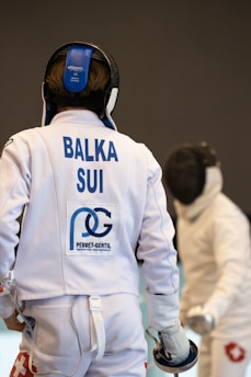 Two fencers are engaged in a bout. The foreground fencer, seen from behind, wears protective gear with a white jacket displaying 'BALKA SUI' and other logos. The fencer in the background faces forward with an obscured face due to their mask. Both are poised with their foils.