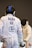 Two fencers are engaged in a bout. The foreground fencer, seen from behind, wears protective gear with a white jacket displaying 'BALKA SUI' and other logos. The fencer in the background faces forward with an obscured face due to their mask. Both are poised with their foils.