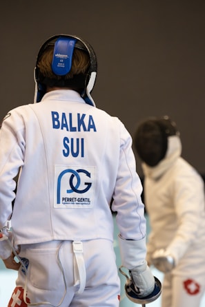 Two fencers are engaged in a bout. The foreground fencer, seen from behind, wears protective gear with a white jacket displaying 'BALKA SUI' and other logos. The fencer in the background faces forward with an obscured face due to their mask. Both are poised with their foils.