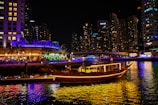 A warmly lit traditional dhow boat cruising past Dubai's illuminated skyline at night.