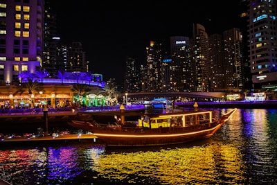 A warmly lit traditional dhow boat cruising past Dubai's illuminated skyline at night.