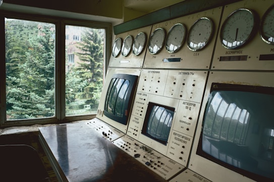A control room with a series of large dials and screens on a console, positioned near a window through which greenery and a building can be seen. The equipment appears aged, with some rust and wear, suggesting it may be from a past era.