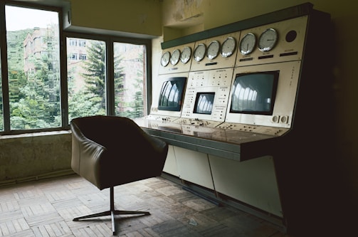 A vintage control room features a large, outdated control panel with numerous dials and screens. The setting is dimly lit, with natural light filtering through large windows showing a view of lush green trees and a distant building. An empty, dark brown swivel chair sits in front of the control panel on a parquet floor.