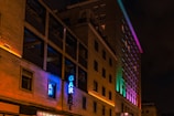 A multi-story building is lit up with colorful neon lights, casting vibrant hues along its facade. The word 'GARAGE' is prominently displayed in blue neon on the side of the building. The structure features rectangular windows, some of which are open, and the lighting creates a vivid, lively effect against the night sky.