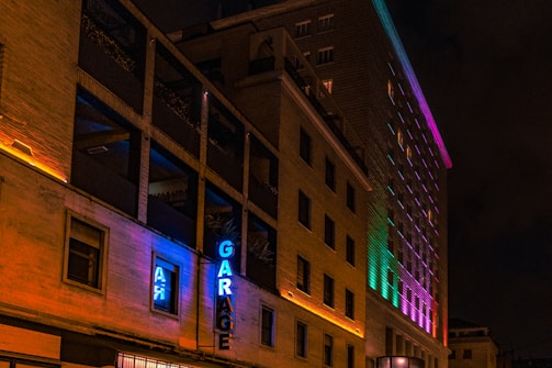 A multi-story building is lit up with colorful neon lights, casting vibrant hues along its facade. The word 'GARAGE' is prominently displayed in blue neon on the side of the building. The structure features rectangular windows, some of which are open, and the lighting creates a vivid, lively effect against the night sky.