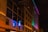 A multi-story building is lit up with colorful neon lights, casting vibrant hues along its facade. The word 'GARAGE' is prominently displayed in blue neon on the side of the building. The structure features rectangular windows, some of which are open, and the lighting creates a vivid, lively effect against the night sky.