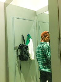 A person in a brown cap is standing in front of a mirror in a fitting room, partially visible, wearing a green plaid shirt. A black backpack and a white and green garment are hanging on the door behind them.