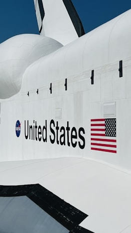 A portion of a space shuttle with the NASA logo and the words 'United States' prominently displayed. The American flag is positioned next to the text on the white surface of the shuttle.