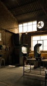 An industrial workshop with brick walls and a high ceiling. The space is lit by natural light coming through large windows, highlighting metal ventilation pipes and workbenches. Some tools and machinery are visible and organized around the room.